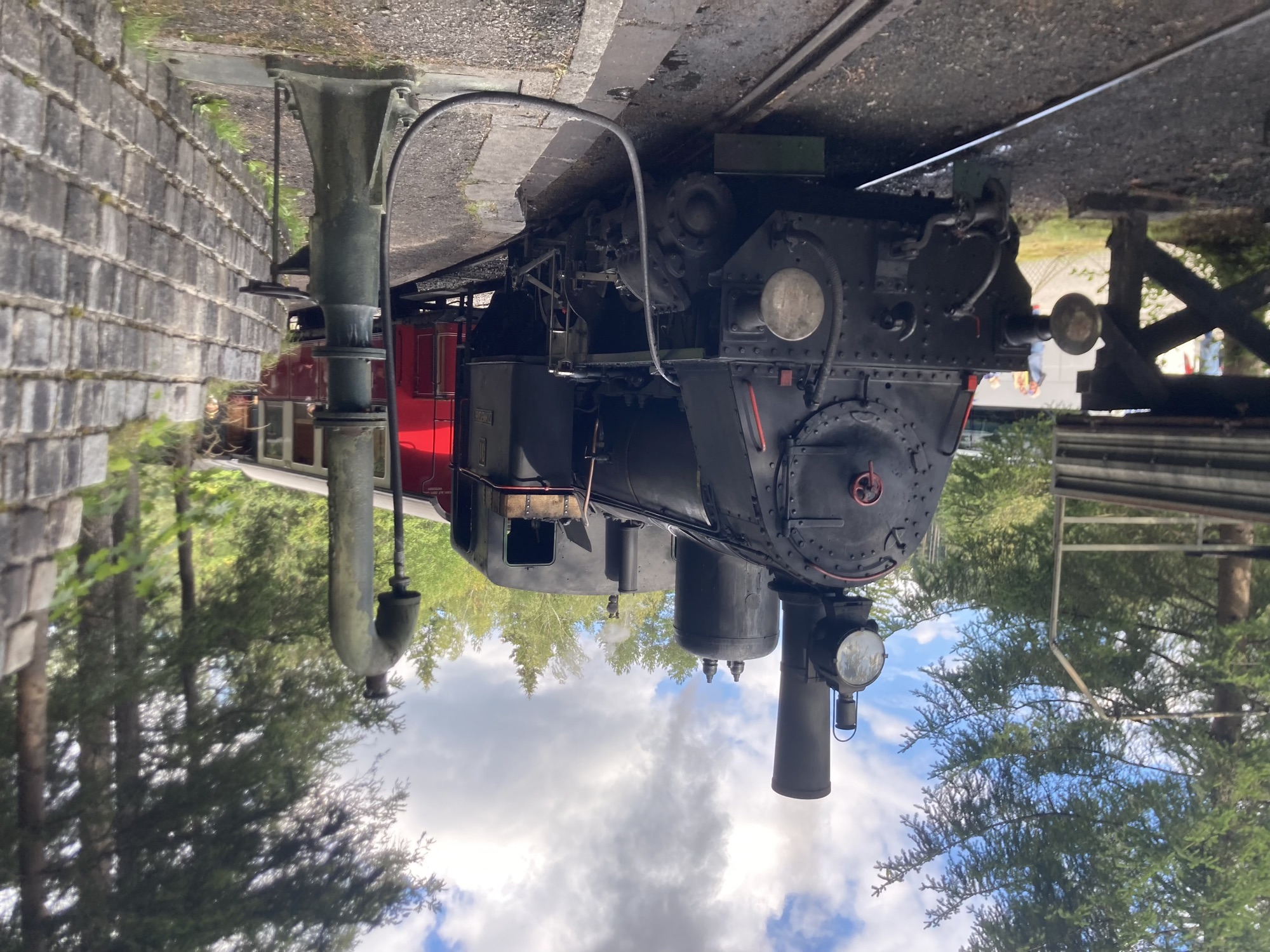 schwarze Dampflokomotive in der Station, von einer Pumpe auf dem Bahnsteig führt ein Wasserschlauch zur Lok, aus den Chimneys strömt grauer Rauch zum Himmel