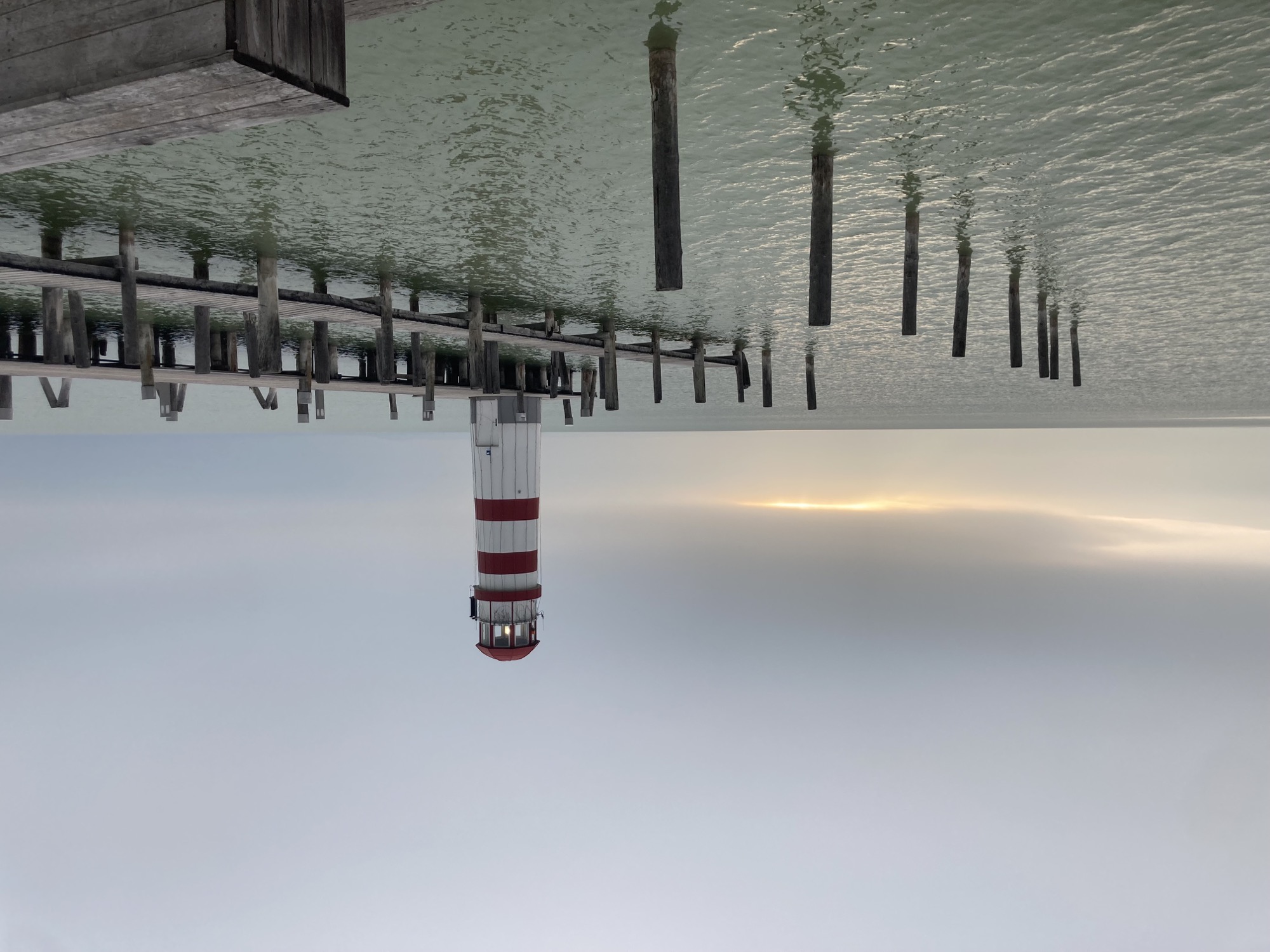 rot-weiß gestreifter Leuchtturm am Ende eines Holzstegs, hinter dem Leuchtturm kämpft sich ein Sonnenstrahl durch die grauen Wolken