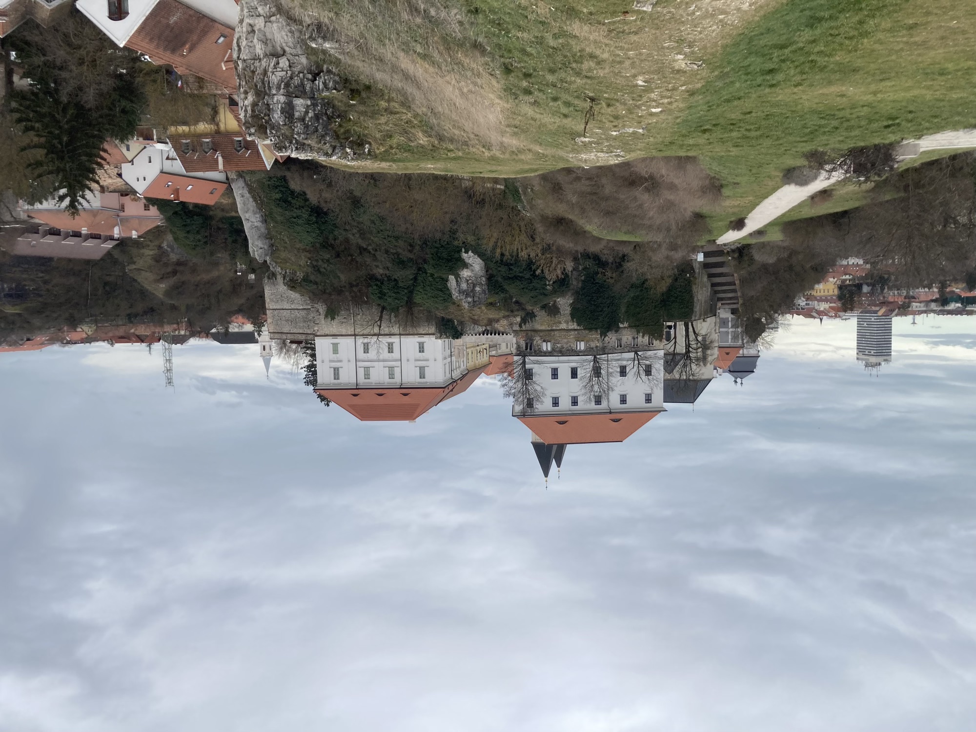 seitliche Ansicht der Burg, vorne ein grüner Weg, der rechts felsig nach unten abfällt, weiter hinten zwei weiße Gebäude über den Burgmauern, darüber sind die Spitzen zweier Kirchtürme zu sehen