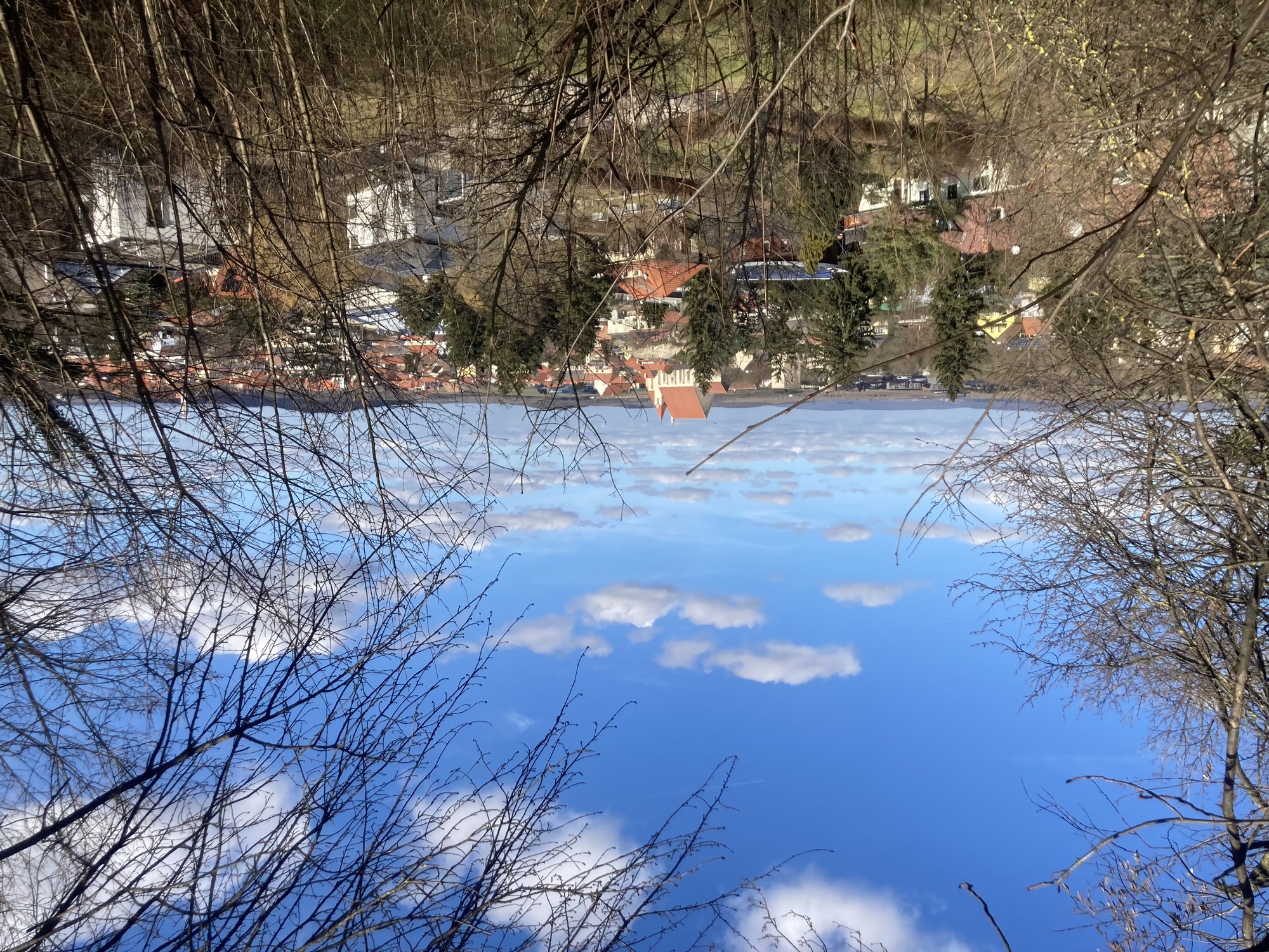 durch kahle Bäume und Sträucher Blick auf eine Ortschaft mit einer Kirche erhöht im Zentrum, die Kirche hat eine ungewöhnliche Form mit einem sehr hohen dreieckigen Dach, das von den Türmen kaum überragt wird