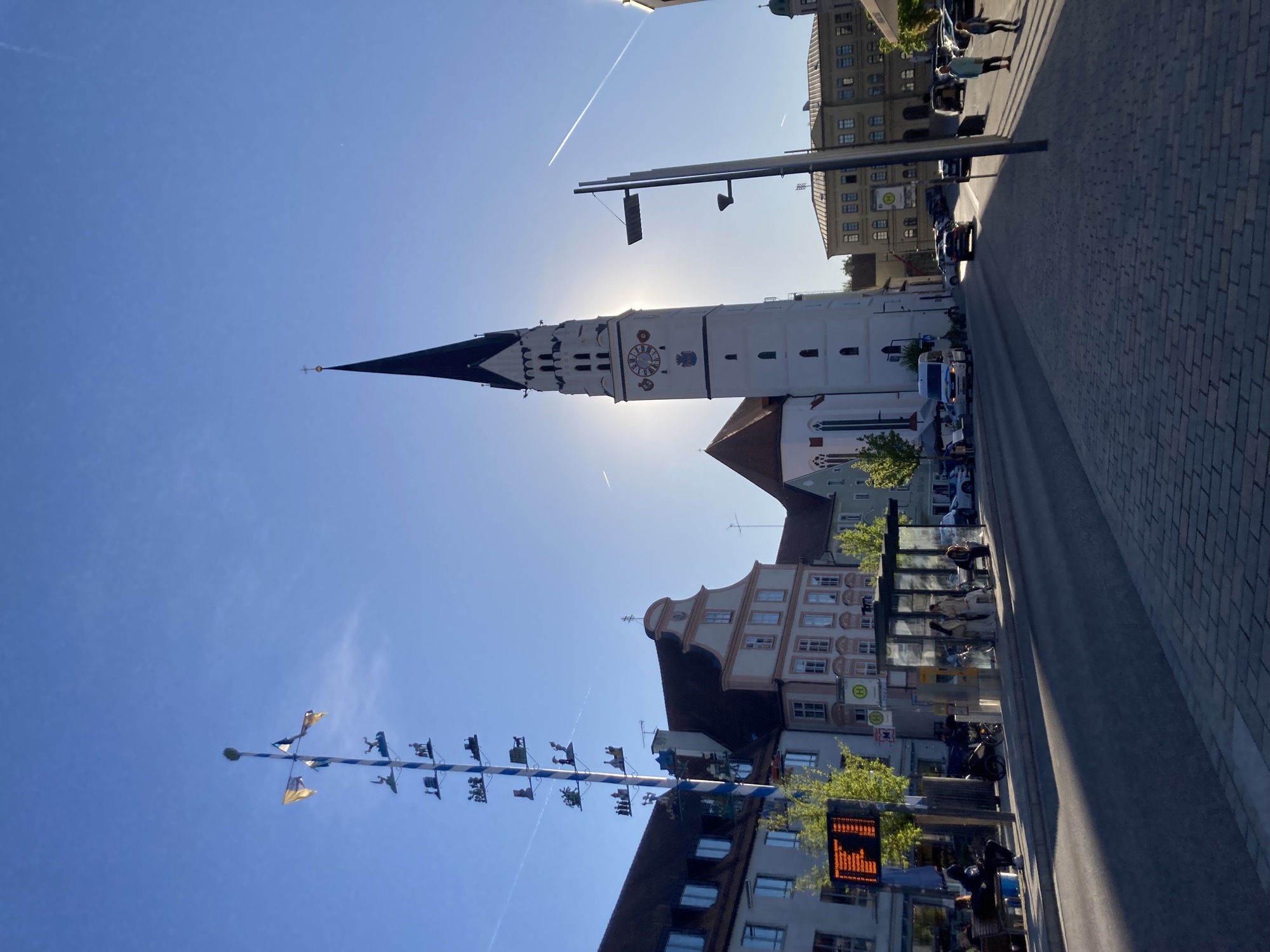 Hauptplatz mit schlankem weißen Kirchturm, die Sonne steht direkt hinter dem Kirchturm, der Turm wirft einen Schatten über den Platz, links von der Kirche im Vordergrund eine Bushaltestelle und ein bayrischer Zunftbaum